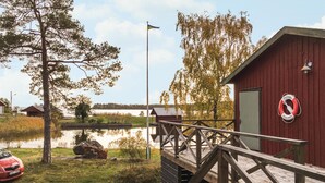 En träbrygga som leder fram till en röd byggnad. På väggen hänger en livboj, det finns en flaggstång och en bil parkerad i närheten.