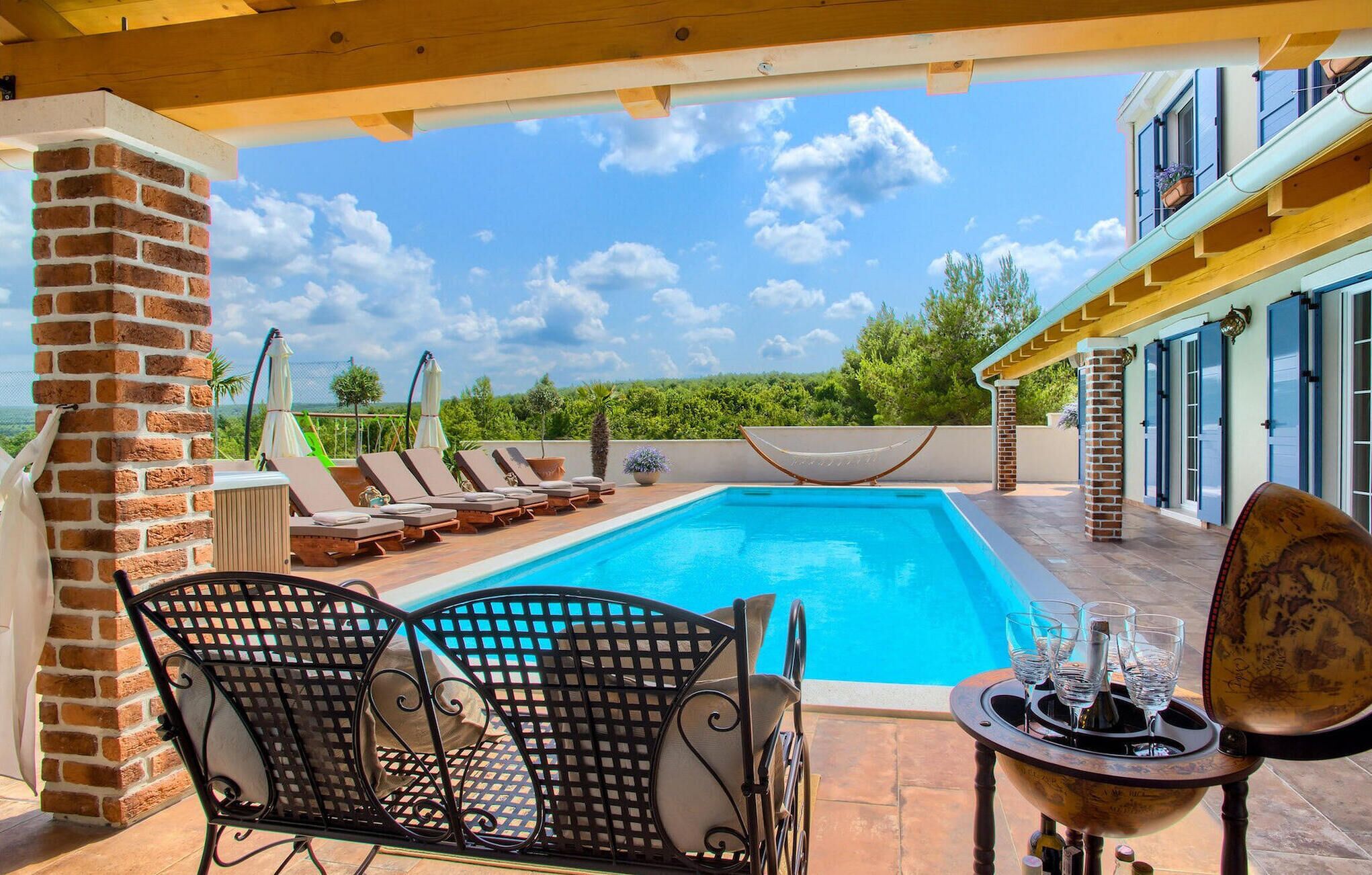 A poolside area with lounge chairs, a table with glasses, and a view of the pool and greenery.