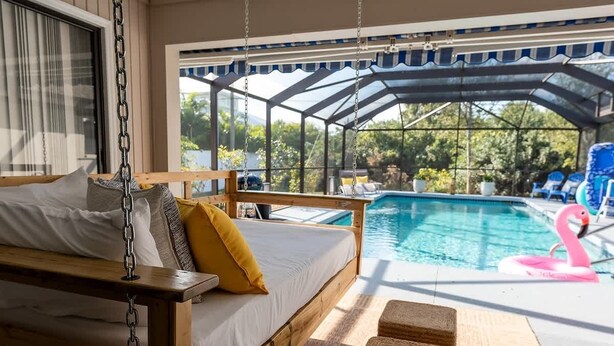 A wooden bed with pillows, a hanging chair, and a view of a pool with a flamingo float. - Heated Pool Oasis w/ Game Room & BBQ Grill (Englewood)