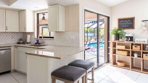 A modern kitchen with white cabinets, a granite countertop, and a bar stool. There is a chalkboard sign that reads "Welcome to Swaying Palms Oasis!" and a coffee station with a sign that says "But First Coffee." - Heated Pool Oasis w/ Game Room & BBQ Grill (Englewood)
