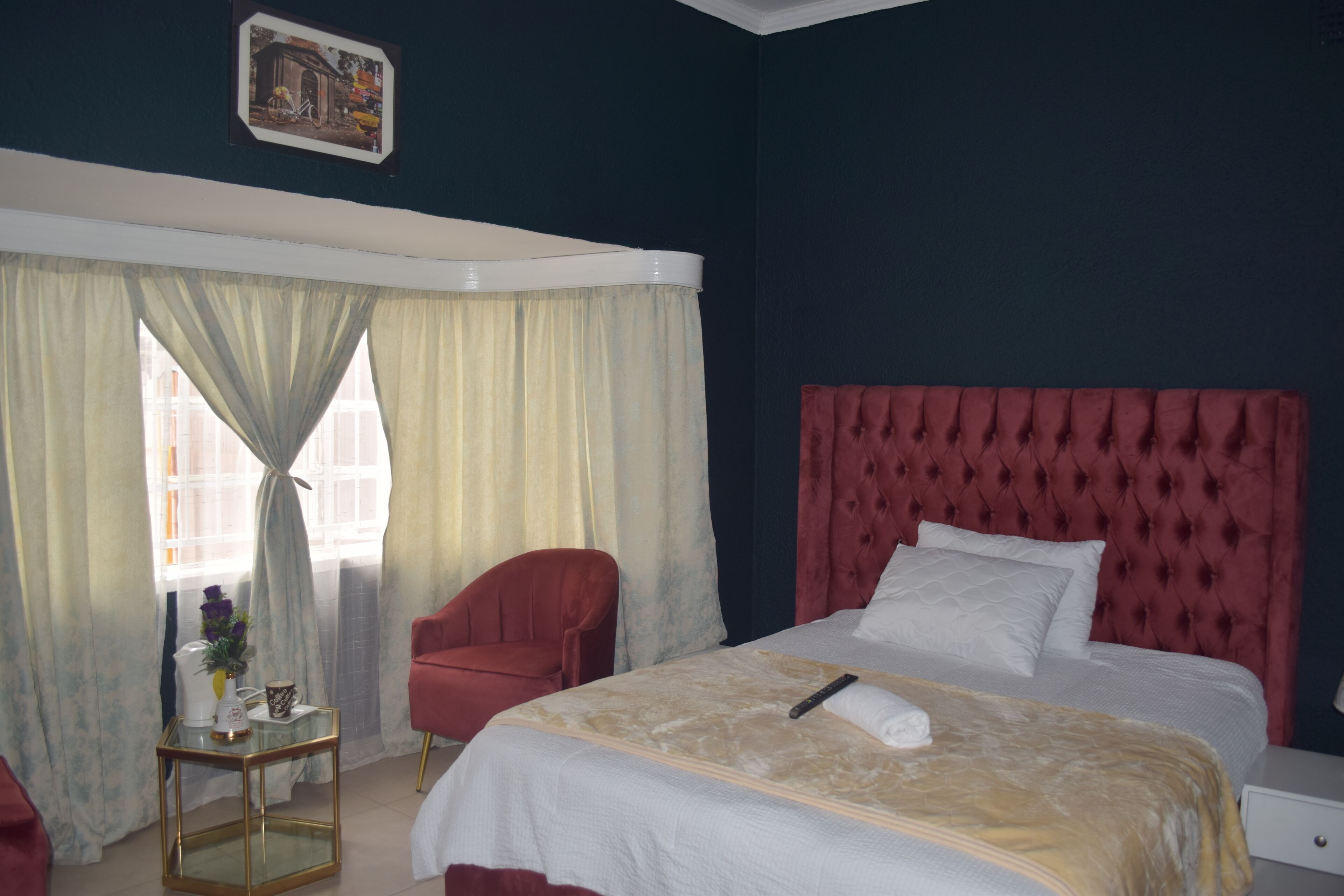 Une chambre d’hôtel avec un grand lit, un fauteuil rouge recouvert de tissu, une petite table avec un vase et une tasse, et une fenêtre avec des rideaux.