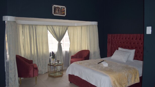 Une chambre d’hôtel avec un grand lit, deux fauteuils rouges, une petite table et une fenêtre avec des rideaux.