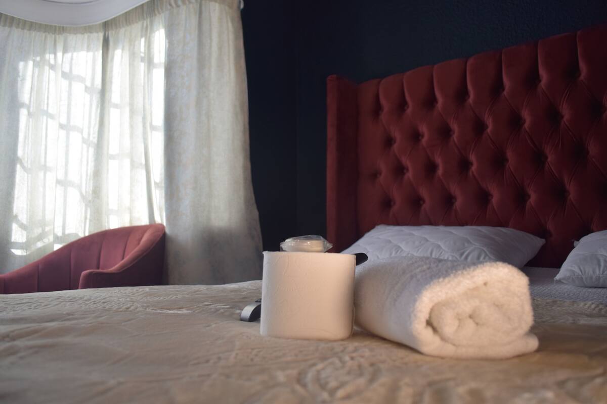Een hotelkamer met een gestoffeerd hoofdeinde, een bed met witte lakens, een opgerolde handdoek, een witte vaas en een rode stoel.