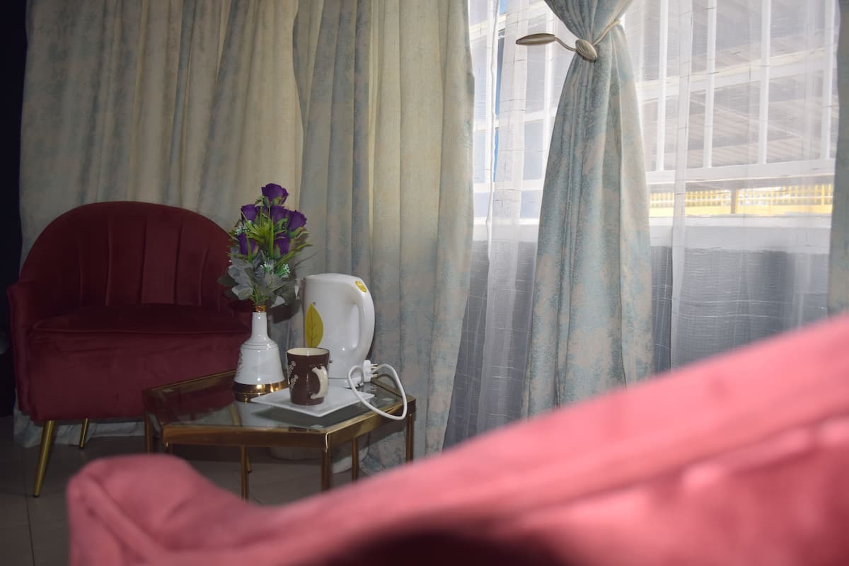 Een kamer met een rode fauteuil, een klein tafeltje met een witte vaas en een bruine mok, en een raam met dunne gordijnen.