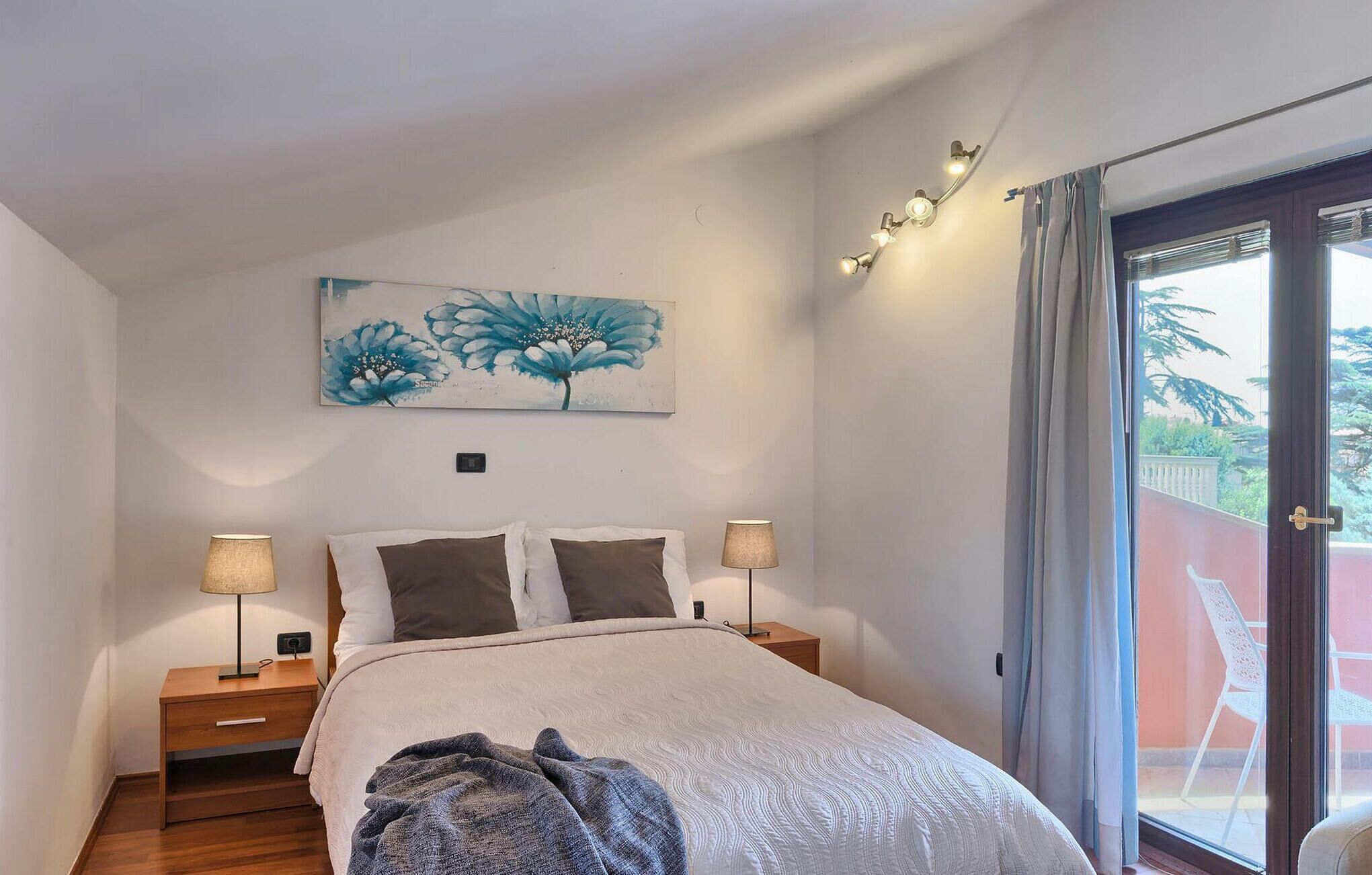 Een slaapkamer met een bed, twee bedlampjes, een nachtkastje, een schilderij en uitzicht op het buitenleven via een schuifdeur.'