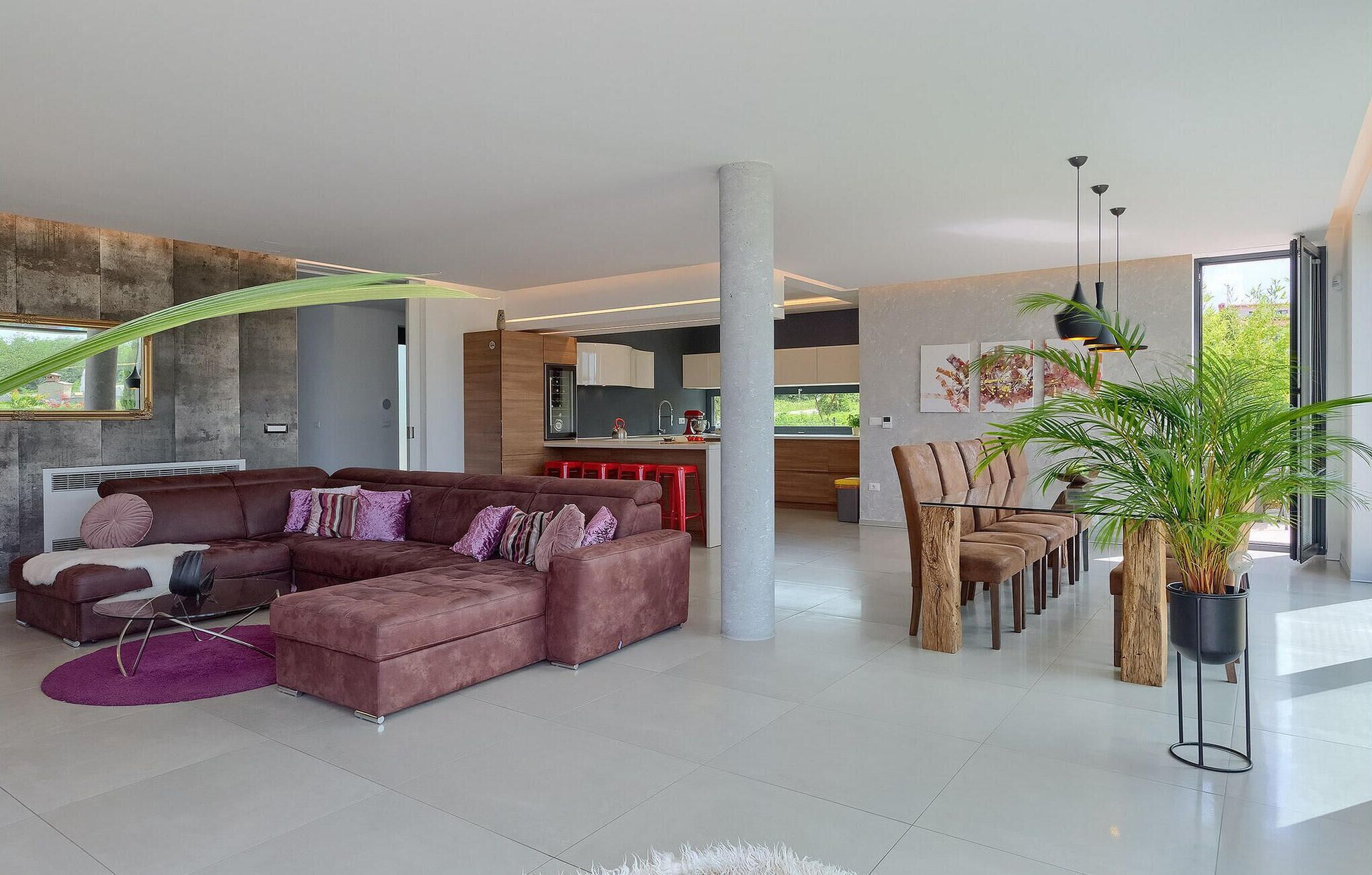 A modern living room with a sectional sofa, a dining area with wooden chairs, a kitchen in the background, and large windows providing natural light.
