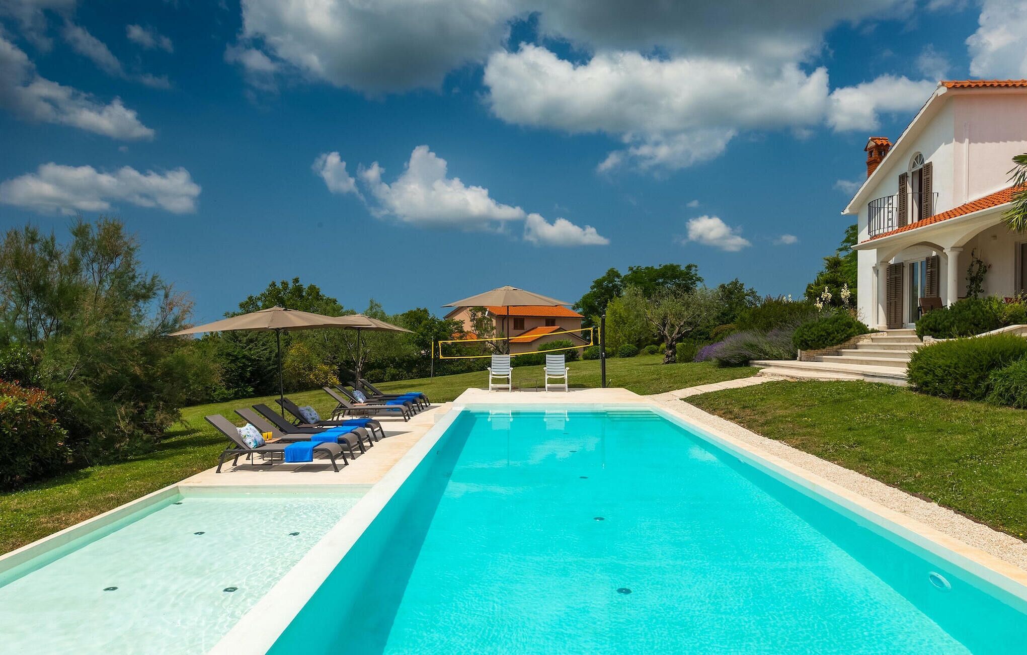 A rectangular swimming pool with lounge chairs and umbrellas, surrounded by greenery and a house with a red-tiled roof.