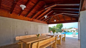 A covered outdoor dining area with a wooden table and benches, a concrete wall, and a view of a swimming pool. - Villa Old School House (Tinjan)