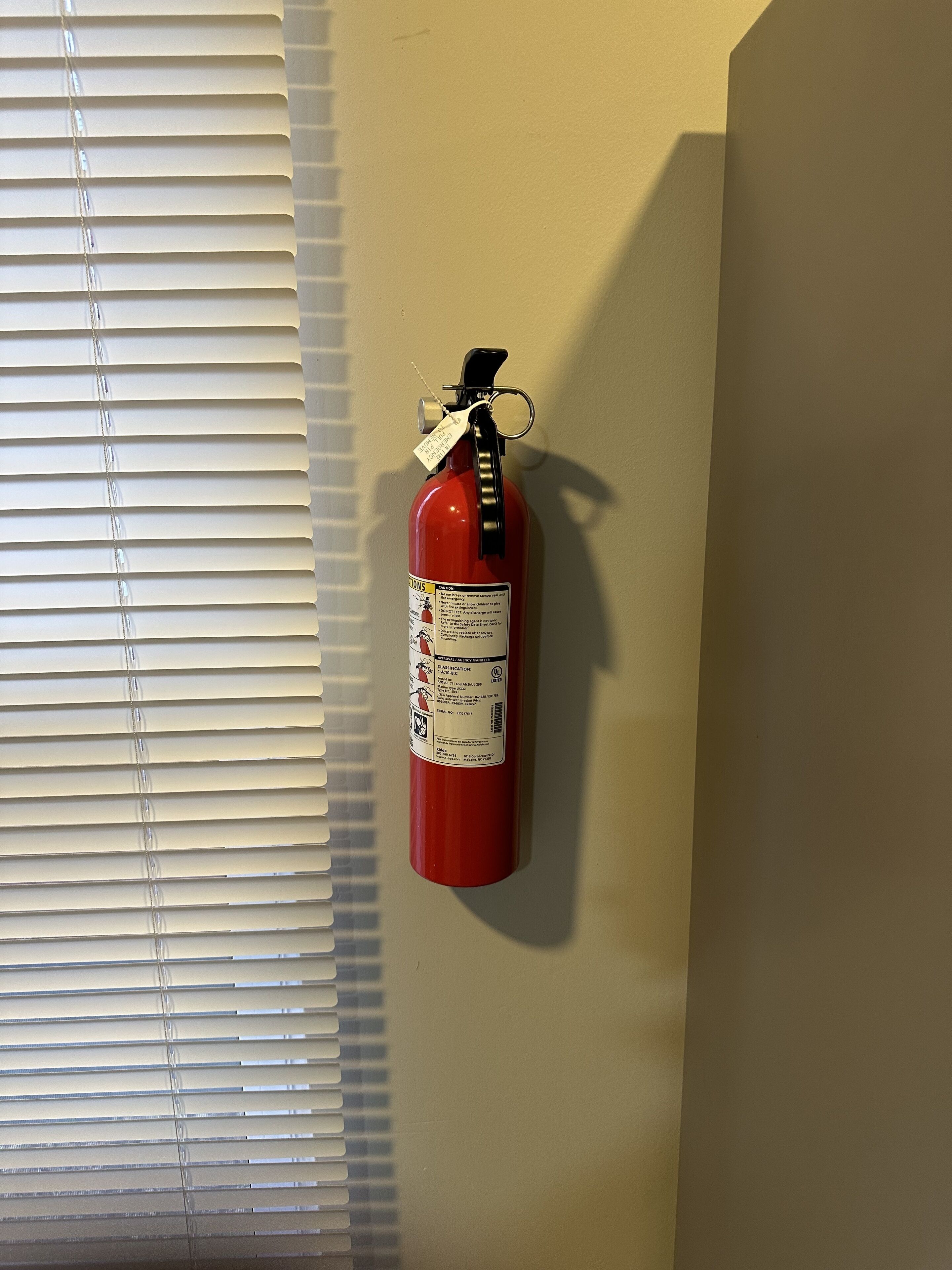 A fire extinguisher mounted on a wall near a window with blinds.
