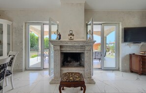 Un salón amplio con chimenea de mármol, un televisor montado en la pared y vistas al exterior a través de puertas de cristal grandes.