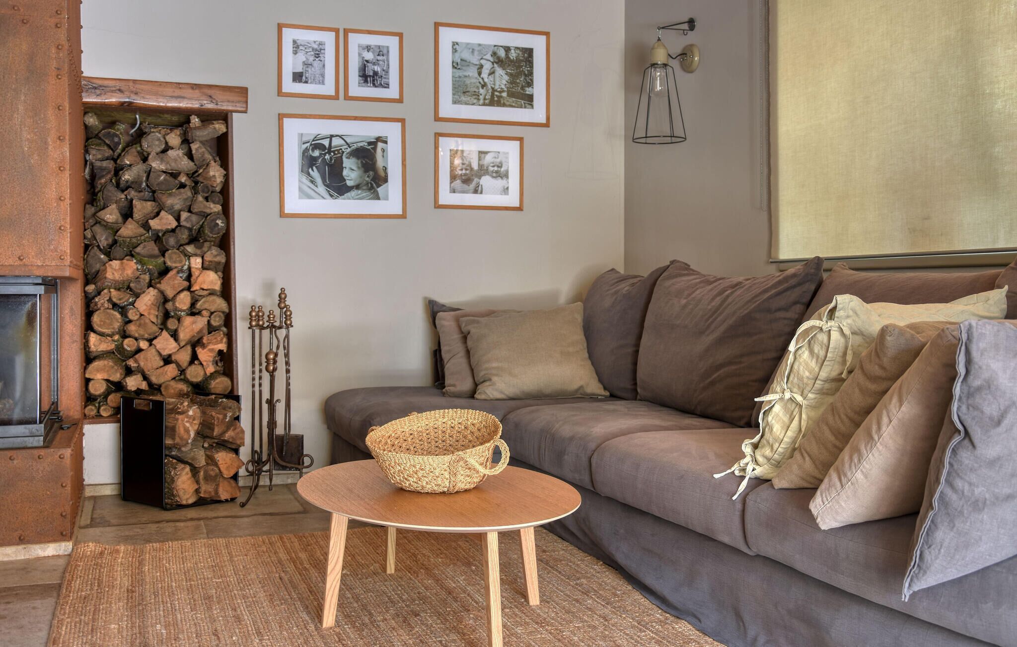 A cozy living room with a grey sofa, a small wooden table with a woven basket, and a fireplace with stacked logs.