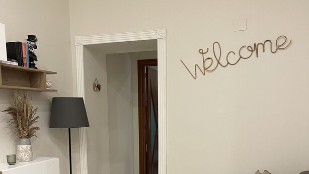 Ein modernes Wohnzimmer mit grauem Sofa, einer Lampe, einem Schild mit der Aufschrift 'Welcome' und einem Flur, der in einen weiteren Raum führt.