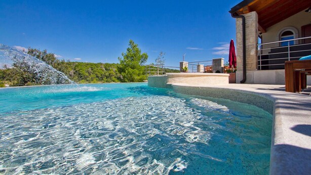 A clear swimming pool with a waterfall feature, surrounded by a stone deck and a wooden pergola. - Gorgeous home in Pula (Pula)