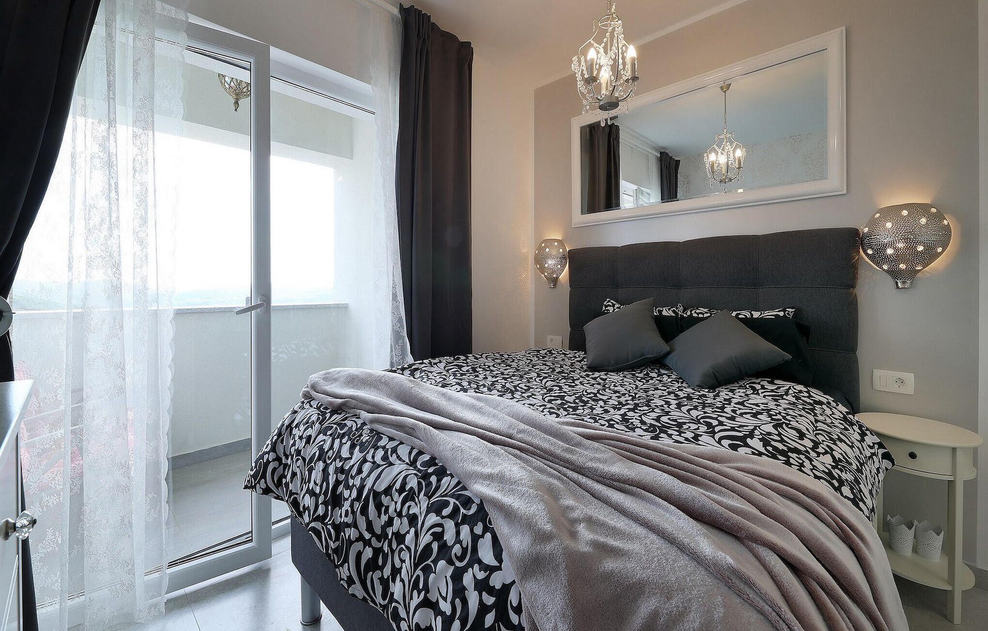 A bedroom with a large bed, a mirror above the bed, two wall sconces, a bedside table, and a view of the outdoors through a sliding door.