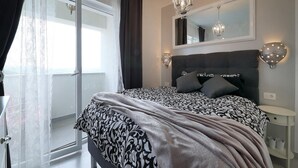 Ein Schlafzimmer mit einem großen Bett, einem Spiegel darüber, zwei Wandleuchten, einem Nachttisch und Blick ins Freie durch eine Schiebetür.
