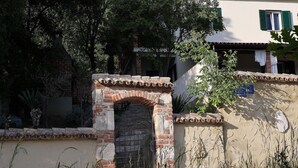 Ein traditionelles Backsteintor mit einer Metalldür, umgeben von Bepflanzung und einem Gebäude mit Ziegeldach und einem blauen Schild.
