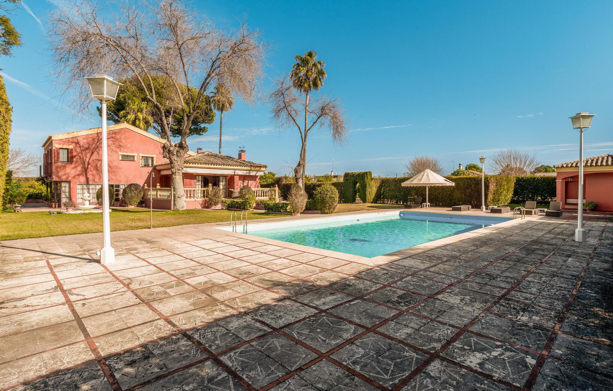 A swimming pool with a tiled deck, surrounded by a house with a red facade, palm trees, and a clear blue sky.