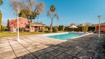 A swimming pool with a tiled deck, surrounded by a house with a red facade, palm trees, and a clear blue sky.