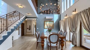 Un vaste espace dédié à la salle à manger, avec une table et des chaises en bois, un escalier doté d’une rampe et une grande fenêtre ornée de rideaux.