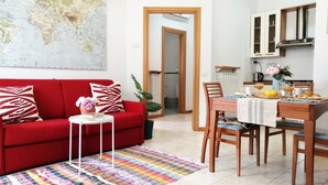 A living room with a red sofa, a small table with a plant, and a colorful rug. There is a dining area with wooden chairs and a table set with a tray of food and drinks.
