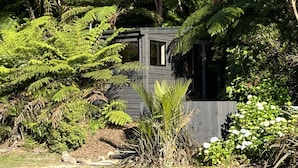 Een klein houten huisje, omringd door weelderige begroeiing, waaronder varens en bloeiende planten.