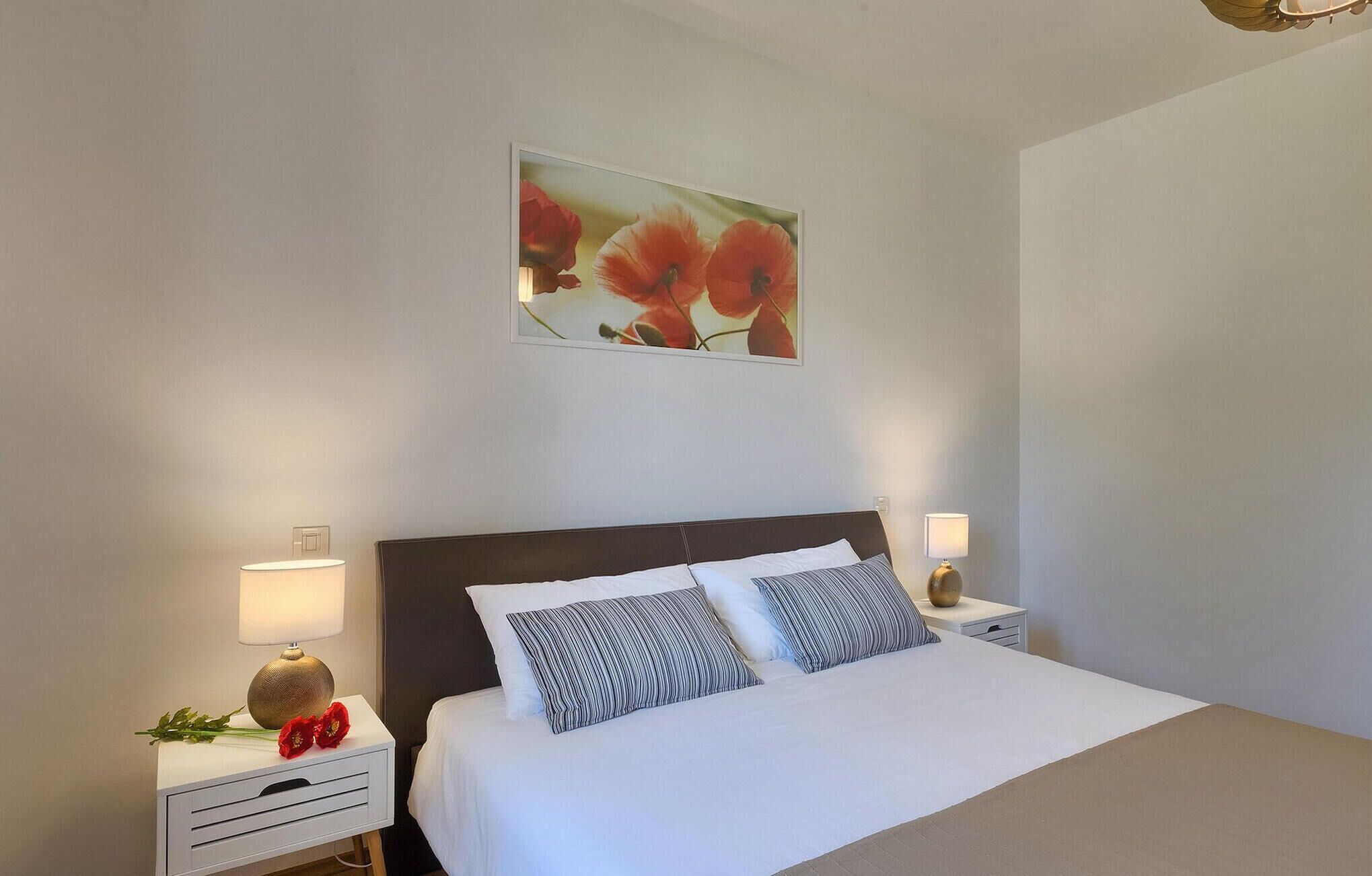 Un dormitorio ordenado con una cama grande, dos mesitas de noche con lámparas y una obra de arte floral enmarcada sobre la cama.