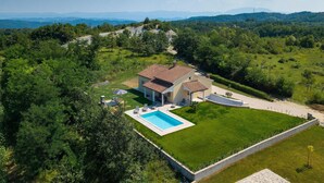 A two-story house with a swimming pool, surrounded by lush greenery and hills in the background.