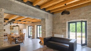 Amplia sala de estar con pared de piedra, vigas de madera, sofá de cuero negro y zona de comedor con sillas y mesa de madera.