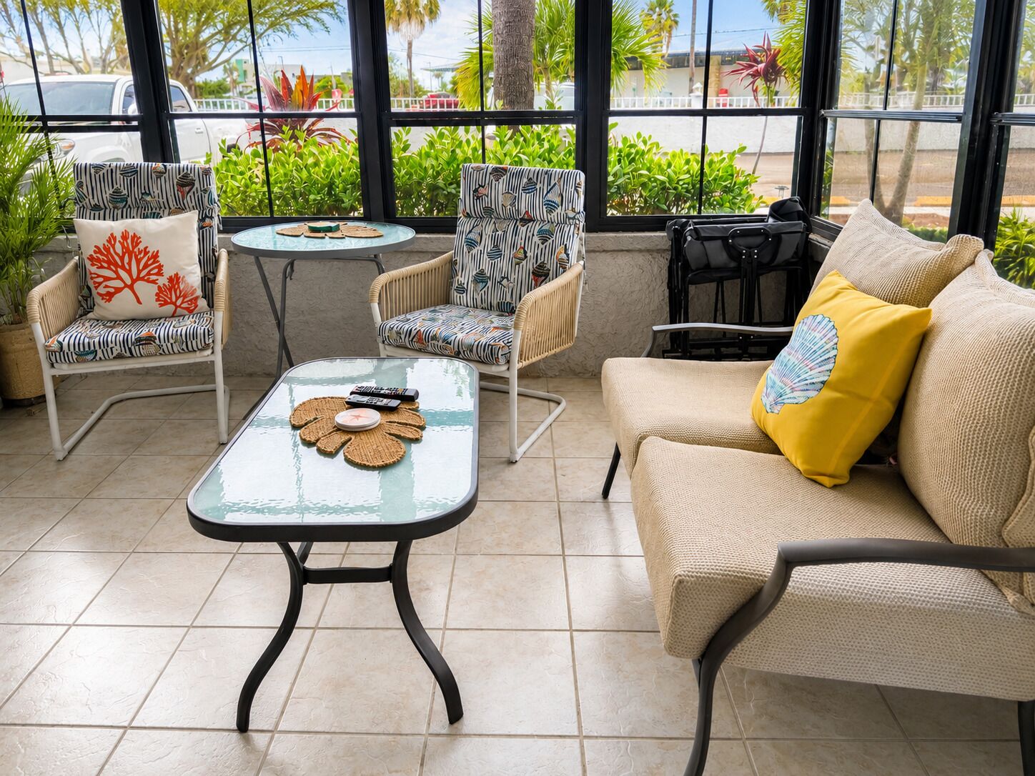 A sunlit patio with wicker furniture, a glass table, and a view of palm trees and parked cars.