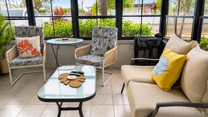 A sunlit patio with wicker furniture, a glass table, and a view of palm trees and parked cars. - Beachy Boho. August specials Waterfront Building 1st Floor, 1 Block to Beach (Madeira Beach)