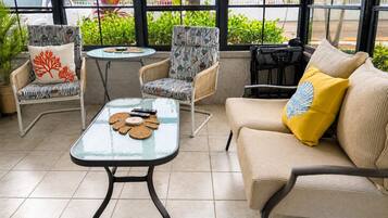 A sunlit patio with wicker furniture, a glass table, and a view of palm trees and parked cars.