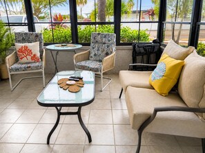 A sunlit patio with wicker furniture, a glass table, and a view of palm trees and parked cars.
