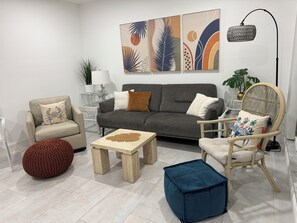 A modern living room with a grey sofa, a wooden coffee table, a red pouf, a blue ottoman, and a wicker chair. - The Beachy Boho. August specials Waterfront Building 1st Floor, Beach Items. (Madeira Beach)