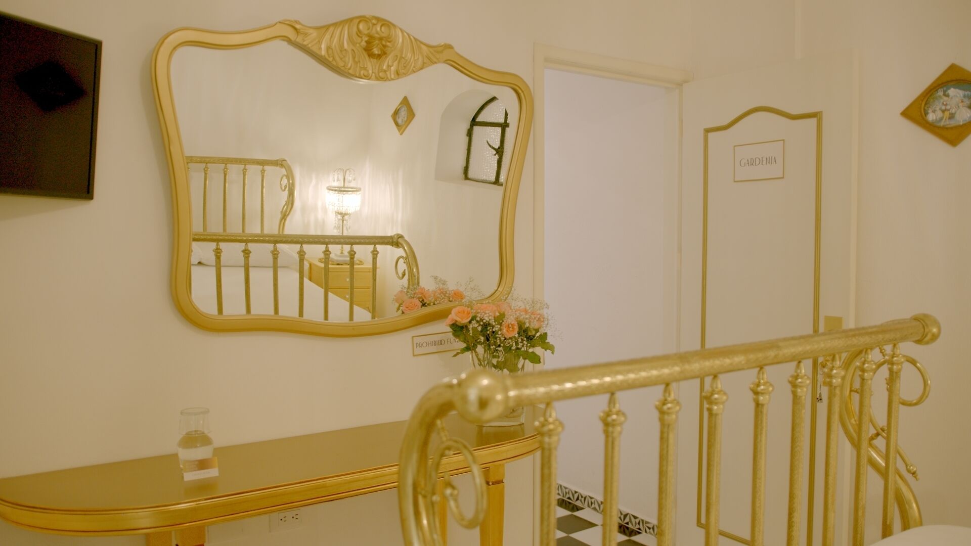 Quarto com um espelho grande e decorativo, uma mesinha de cabeceira com uma garrafa, um arranjo de flores e uma placa com a inscrição 'PROIBIDO ACENDER CHAMA'.