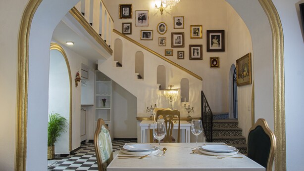 Un comedor con suelo a cuadros, una mesa blanca dispuesta para dos personas y una escalera con candelabro.