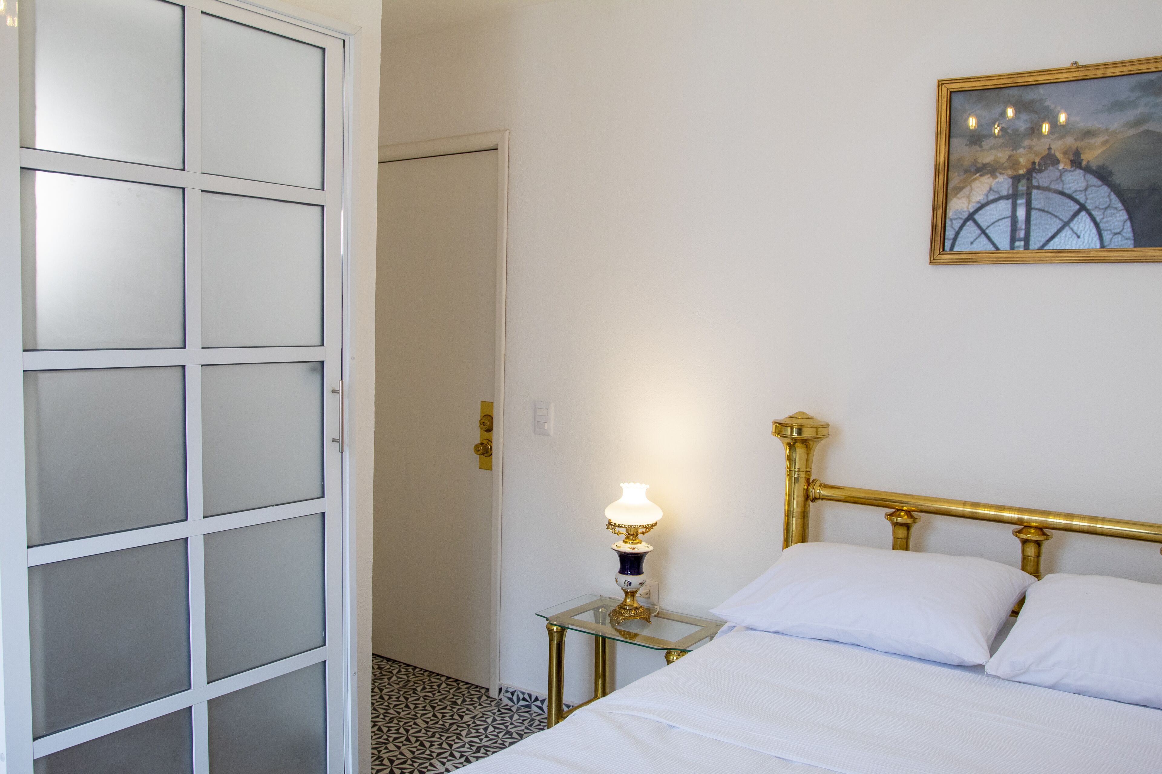 Una habitación con cama, mesita de noche con lámpara, puerta con paneles de vidrio esmerilado y un cuadro en la pared.