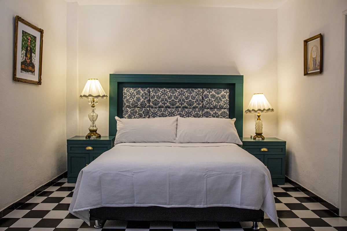 Una cama bien hecha con un cabecero estampado, dos mesitas de noche con lámparas y un suelo a cuadros.