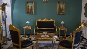 Una habitación con piso a cuadros blanco y negro, un sofá dorado y negro muy decorado, dos sillas a juego, una pequeña mesa redonda con un libro y un vaso, y dos cuadros en paredes verde azulado.