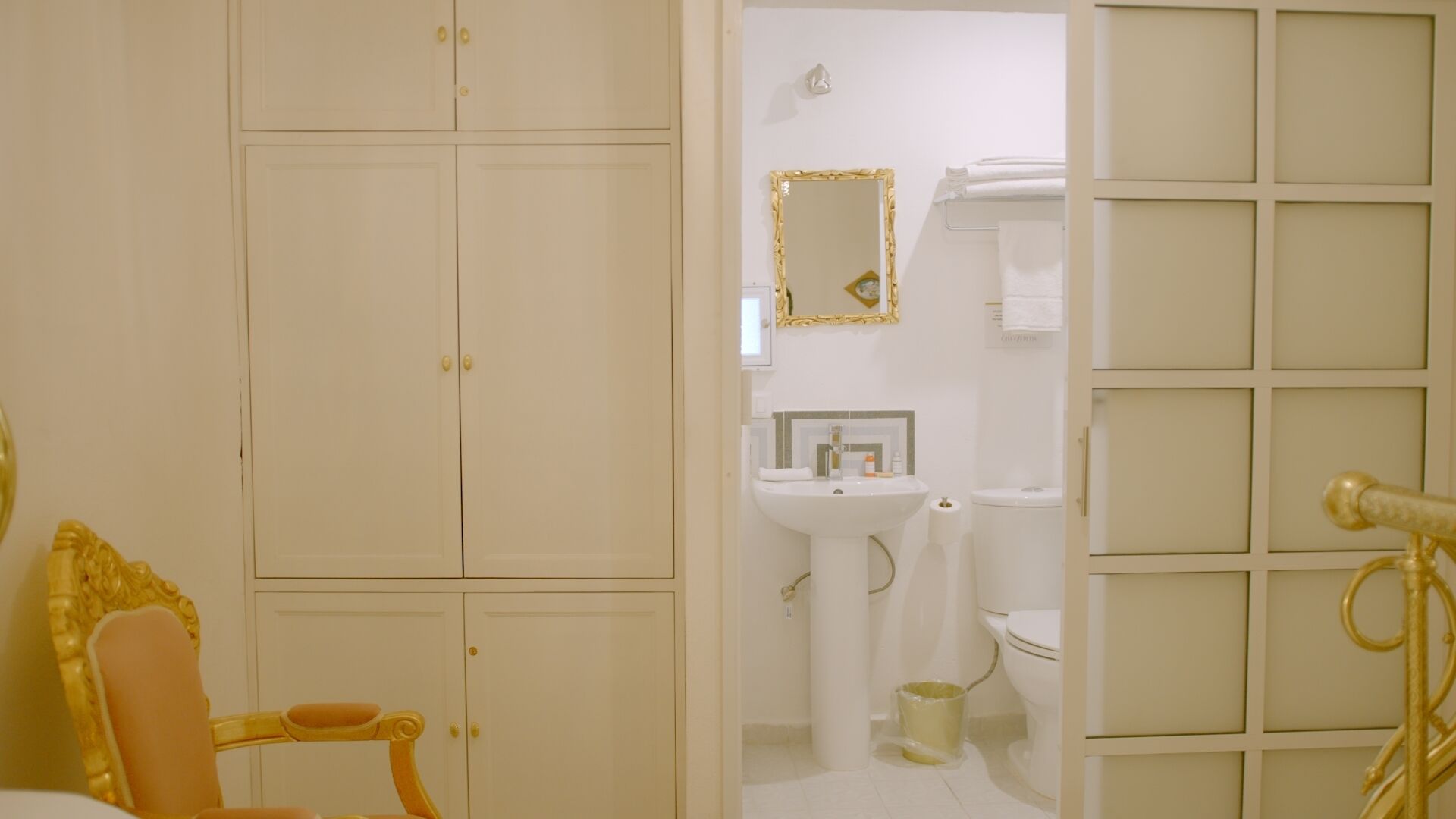 Un baño con lavamanos, inodoro y espejo blancos. A la derecha, hay una silla con estructura dorada y un objeto color dorado.