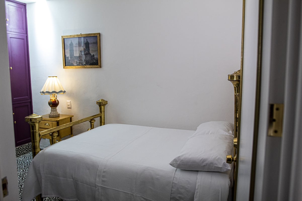 Una cama bien hecha con sábanas blancas, un cabecero con marco dorado y una mesita de noche con una lámpara. Un cuadro enmarcado cuelga de la pared sobre la cama.
