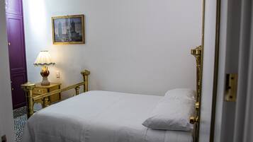 Una cama bien hecha con sábanas blancas, un cabecero con marco dorado y una mesita de noche con una lámpara. Un cuadro enmarcado cuelga de la pared sobre la cama.