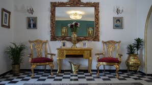 Una habitación con piso a cuadros, dos sillones dorados ornamentados con cojines rojos, un escritorio dorado, un espejo grande con marco dorado y una lámpara de araña.