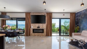 Ein modernes Wohnzimmer mit Kamin, Flachbildfernseher, Essbereich mit Blick ins Freie und Balkon mit Meerblick.