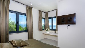 Ein Schlafzimmer mit Bett, Fenster mit Vorhängen, Nachttisch mit Blumenvase und Spiegel über dem Waschbecken.