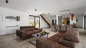 Un salon moderne avec un canapé en cuir marron, une table basse en verre et un coin repas avec des chaises grises. Un escalier en bois mène à un autre niveau.