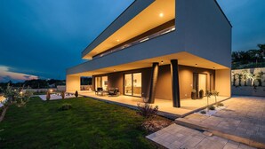 Une maison moderne à deux étages, dotée d’un toit plat, de grandes fenêtres et d’un vaste espace extérieur aménagé en patio.