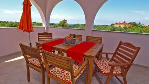 Un tavolo di legno con tovaglie rosse e sedie con cuscini colorati, su un balcone con archi e un ombrellone arancione.