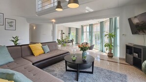 Uma sala de estar moderna com um sofá, mesa de centro e uma televisão. Plantas e uma área de jantar são visíveis ao fundo.