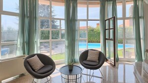 Quarto com janelas amplas, duas cadeiras pretas, uma mesa pequena e vista para uma piscina e vegetação.
