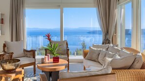 Ein geräumiges Wohnzimmer mit einem großen Fenster und Meerblick. Es gibt einen runden Holztisch mit einer Pflanze, ein Sofa und einen Sessel.
